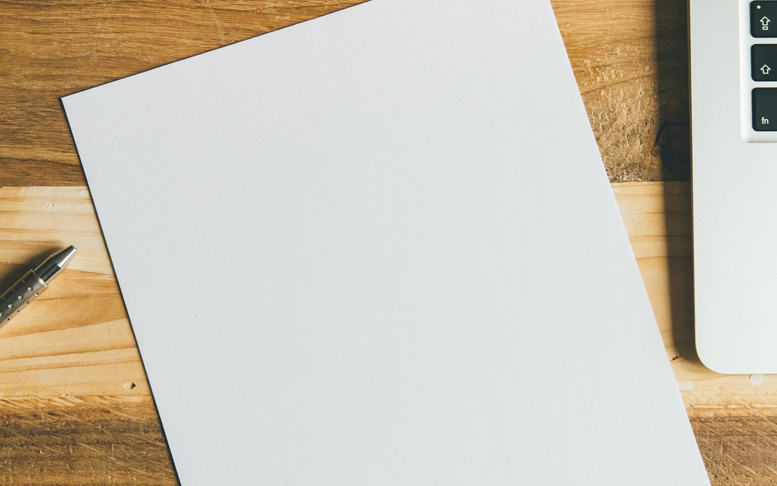 Hoja en blanco y pluma sobre un escritorio de madera junto a una laptop. Foto: Markus Spiske, Unsplash
